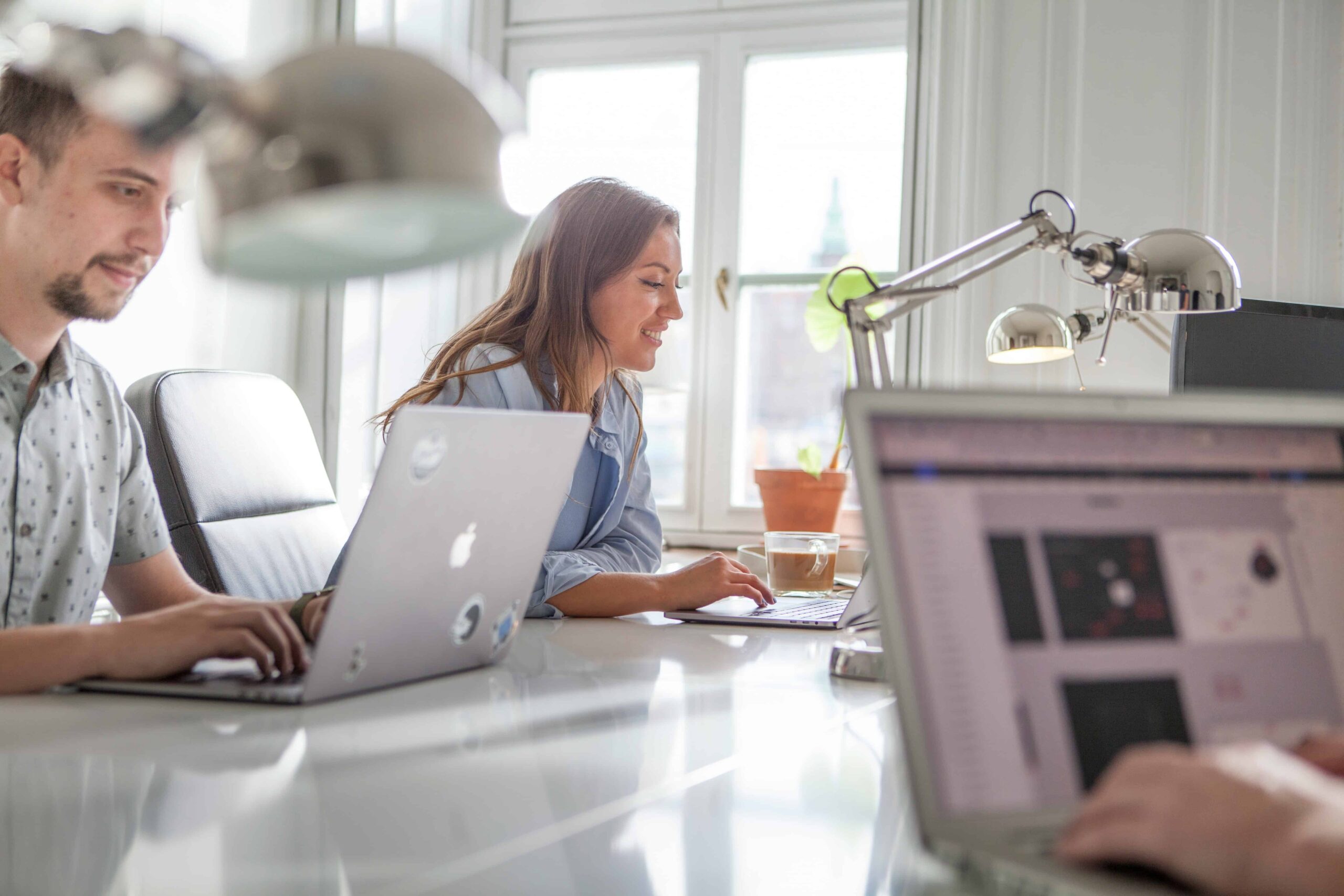 A man and a woman working on laptops in a bright office, representing collaboration, creativity, and what About Now and Every Other Day stands for.